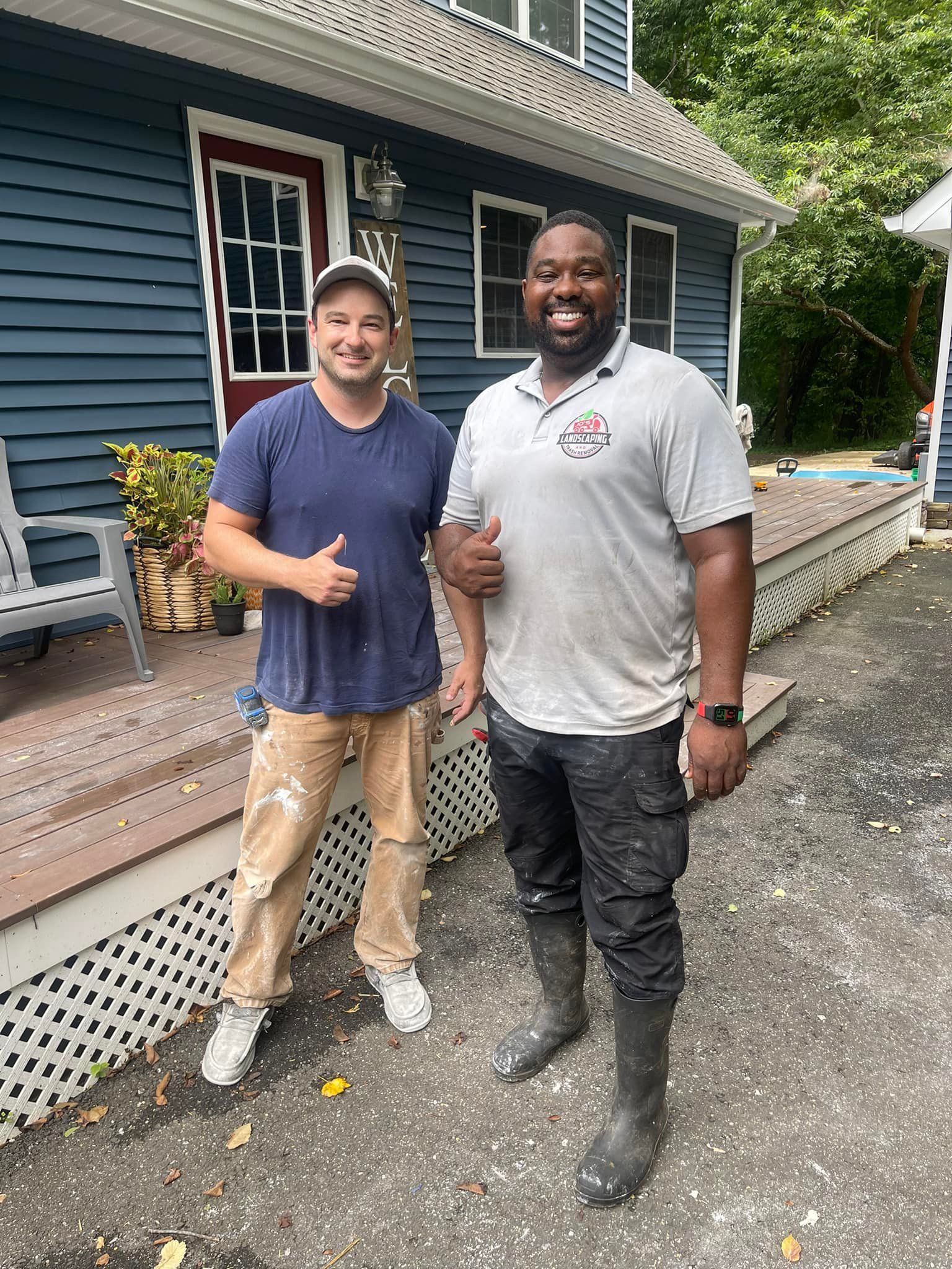 Two people posing with thumbs up in front of a blue house, wearing work clothes and boots on a gravel driveway.