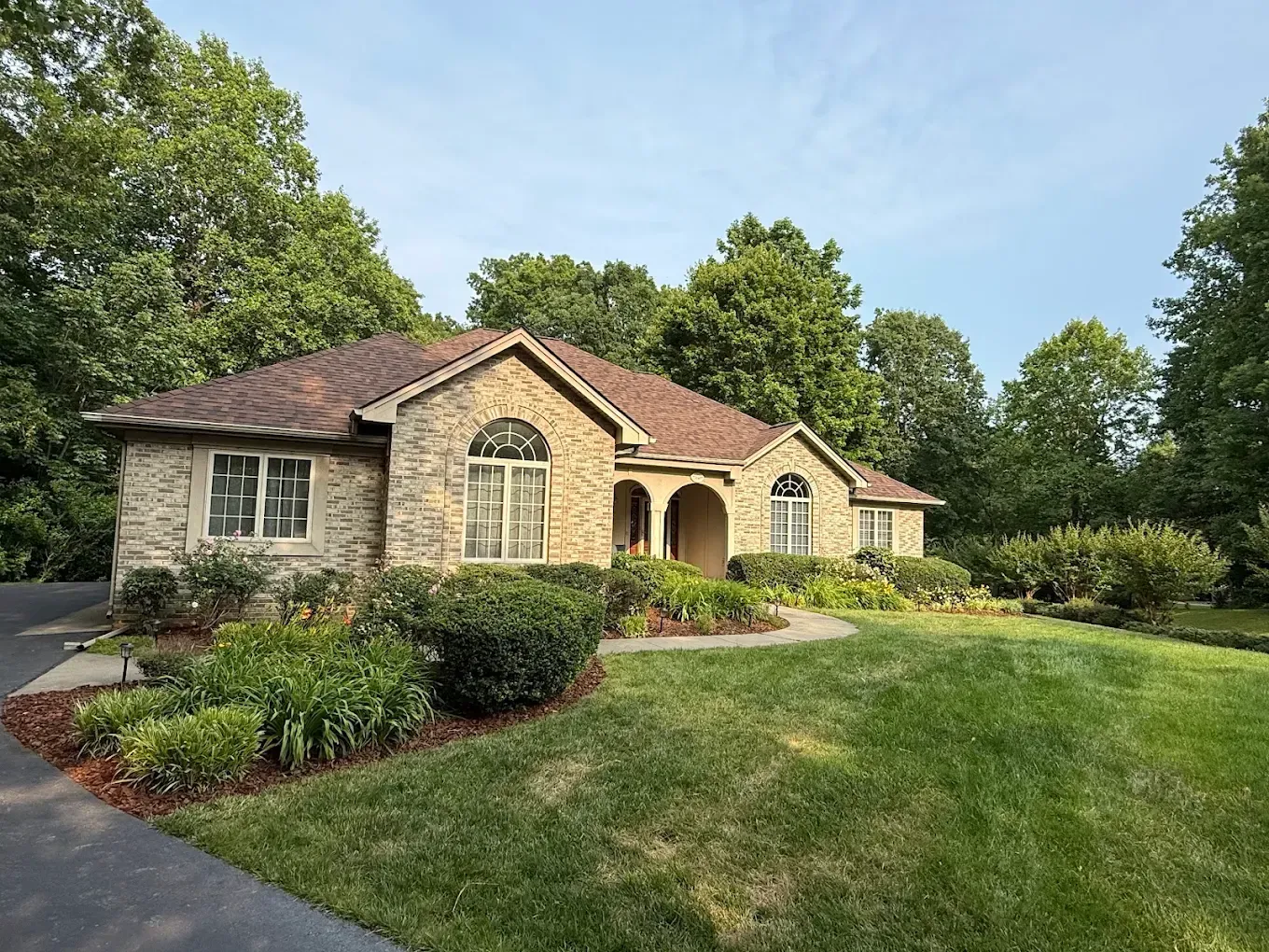A single-story, stone-faced house with a brown shingled roof, surrounded by green trees and a well-manicured lawn.