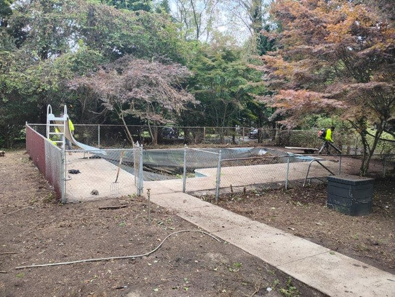 Construction site with concrete footing, fenced-off area, and a worker in a yellow vest among trees.