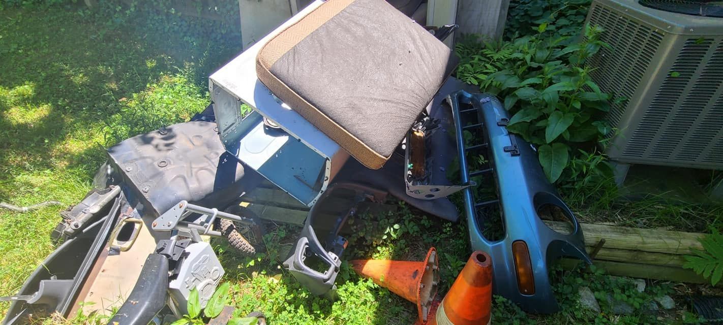 A pile of salvaged car parts, including a bumper and seat cushion, sits on the grass near an outdoor air conditioner.