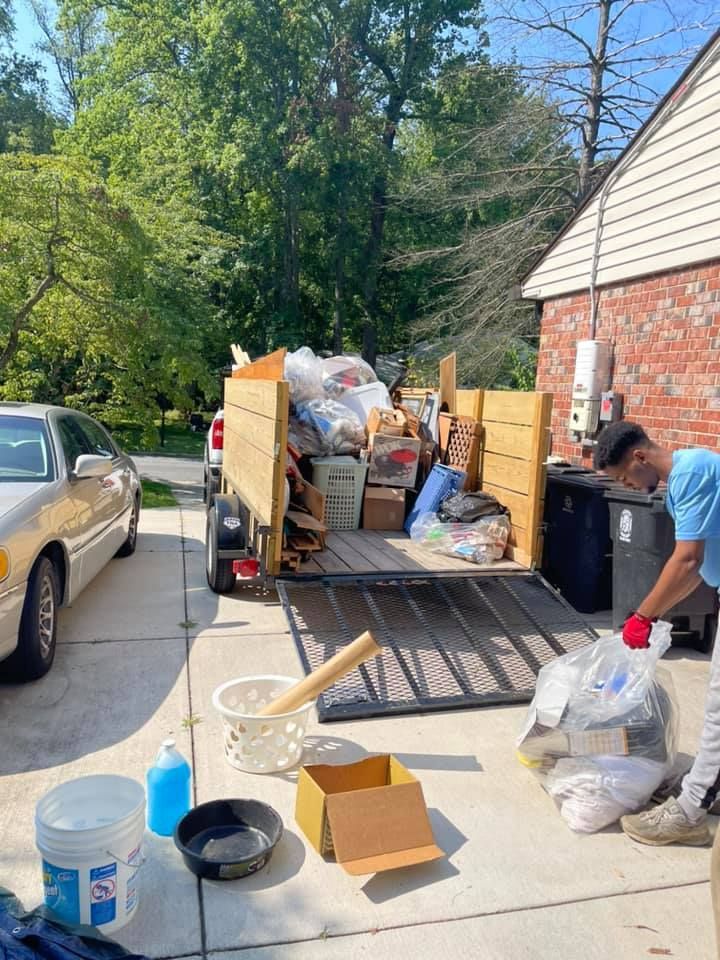 A person loading trash into a utility trailer parked in a driveway next to a parked car and a brick house.