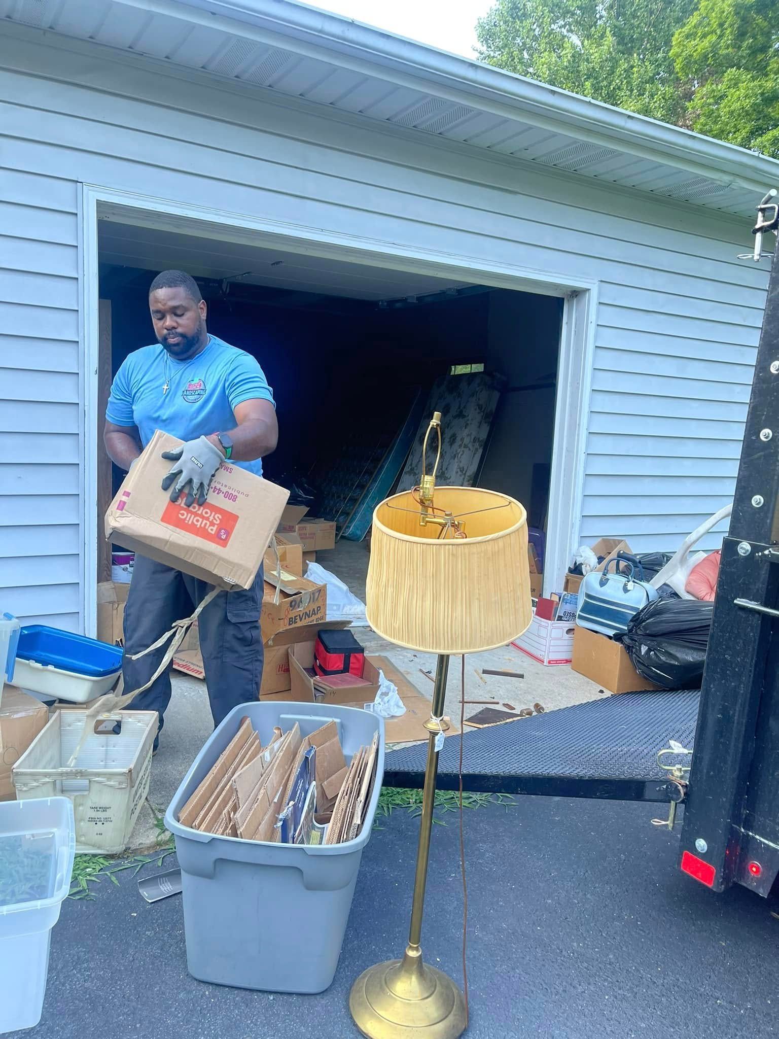A person in a blue shirt holds a cardboard box in front of a garage, with a gold floor lamp in the foreground.