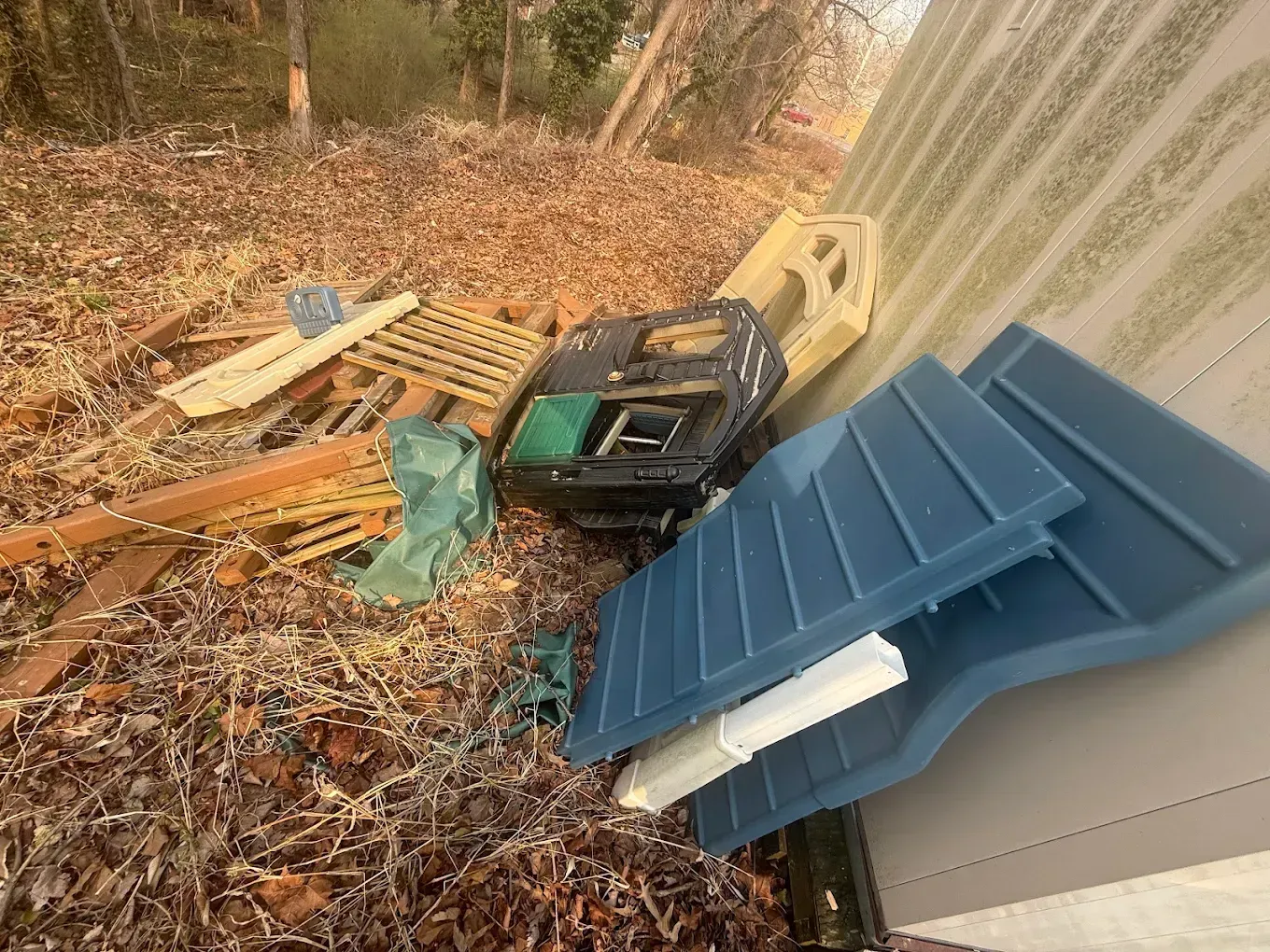 A pile of discarded items, including a blue plastic roof piece, wooden debris, and a black storage bin, sits outdoors.