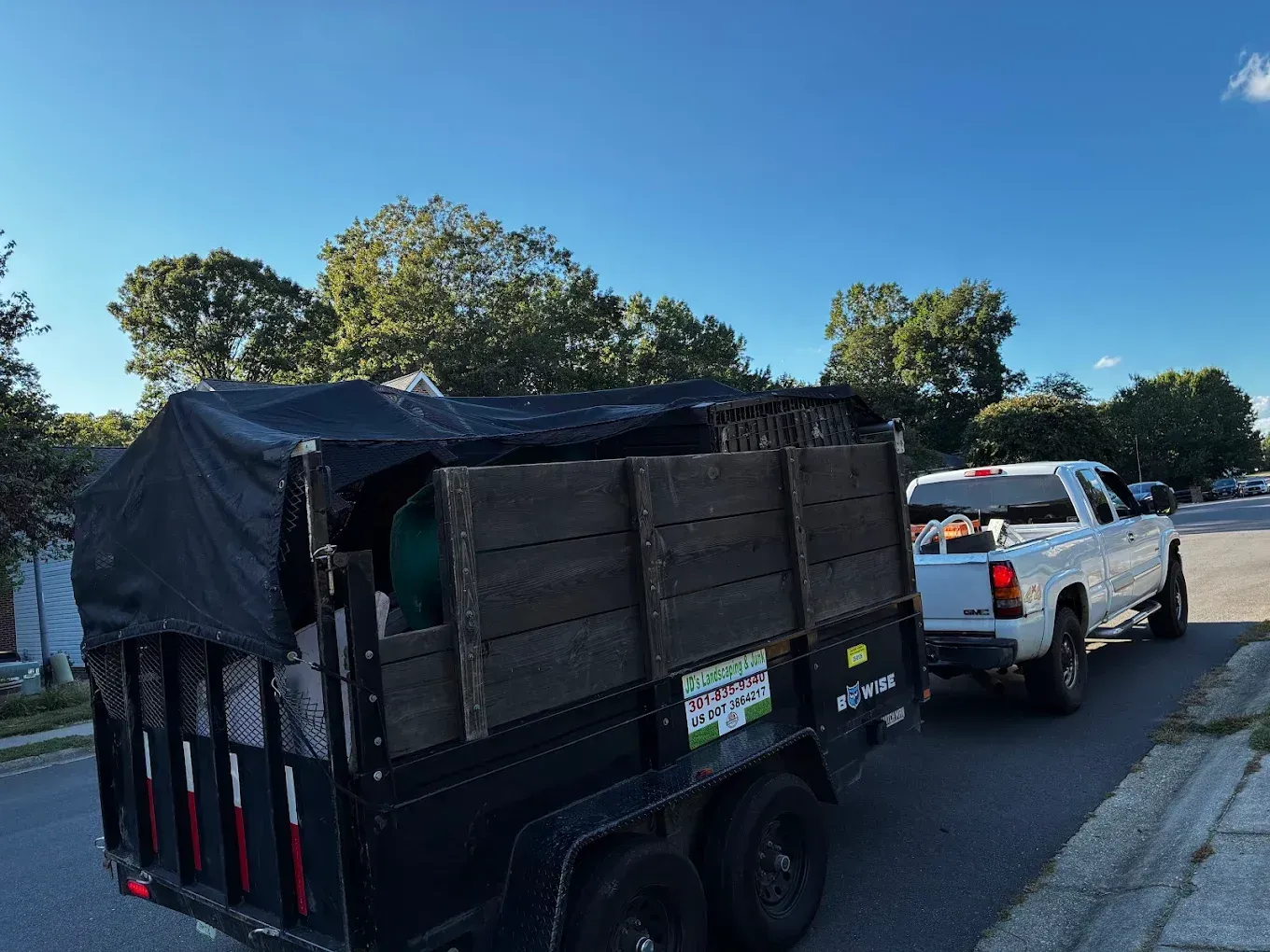 A white pickup truck pulls a black, wooden-sided utility trailer covered with a black tarp down a residential street.
