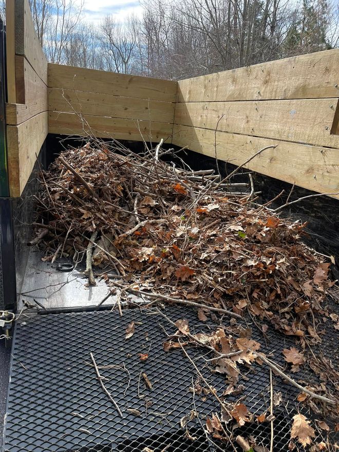 A groundskeeper in a green uniform uses a backpack leaf blower to clear leaves from a park walkway.
