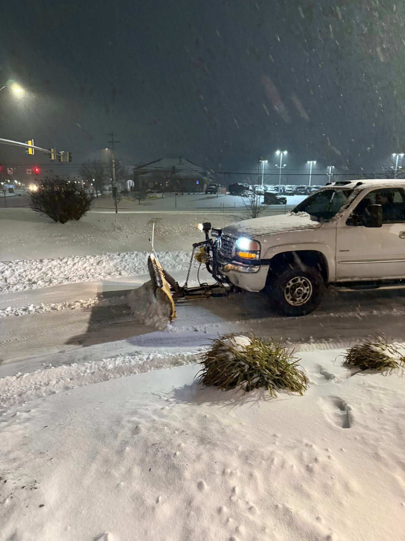 A white pickup truck with an attached snowplow clears snow in a parking lot during a nighttime snowstorm.