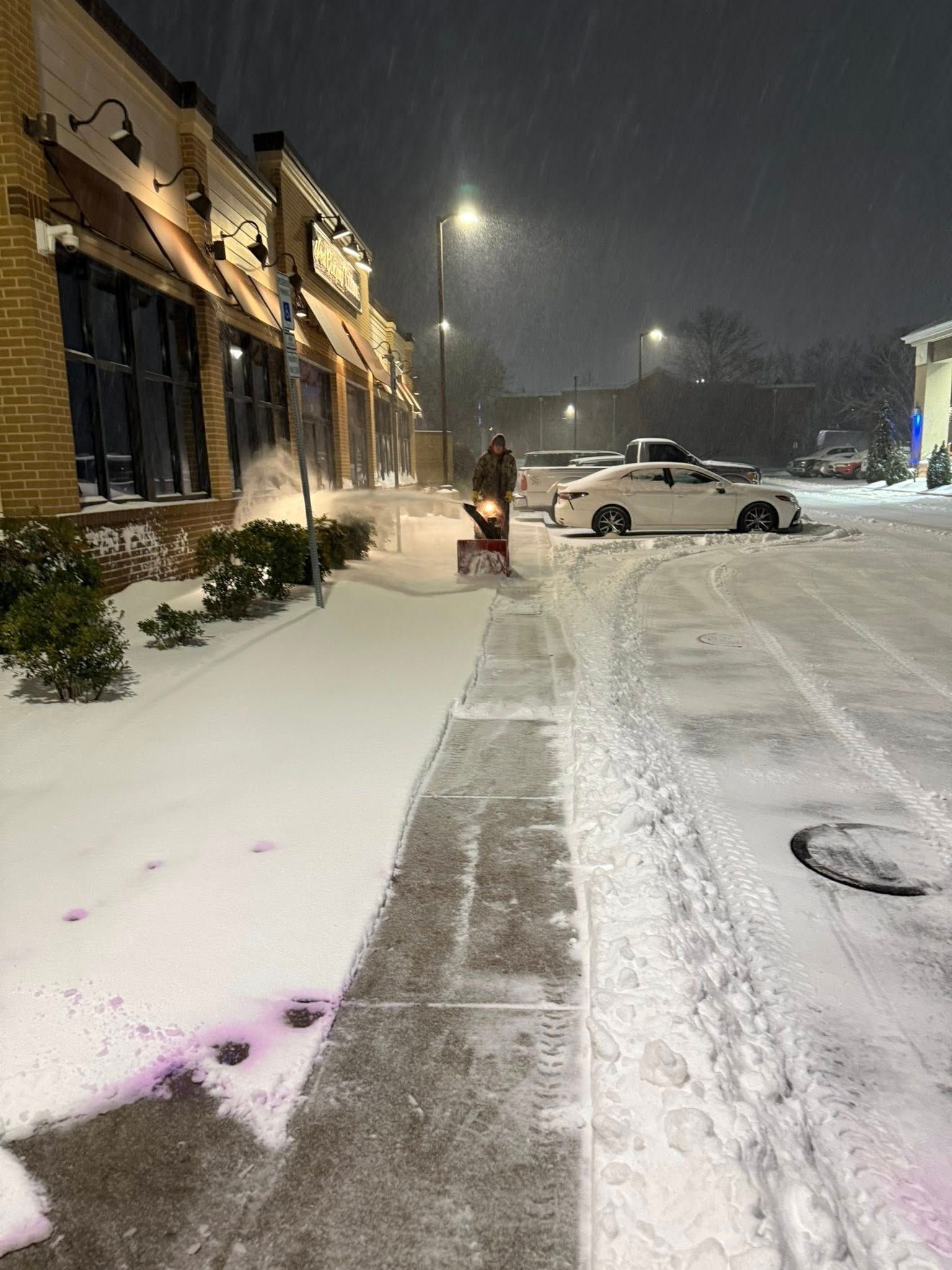A person uses a snowblower to clear a sidewalk in front of a building at night during a snowstorm.