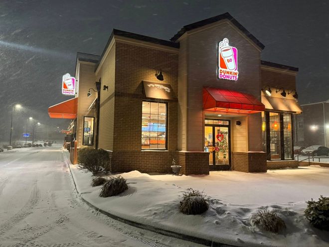 A Dunkin' restaurant at night during a snowfall, with orange awnings and illuminated signs against a snowy parking lot.