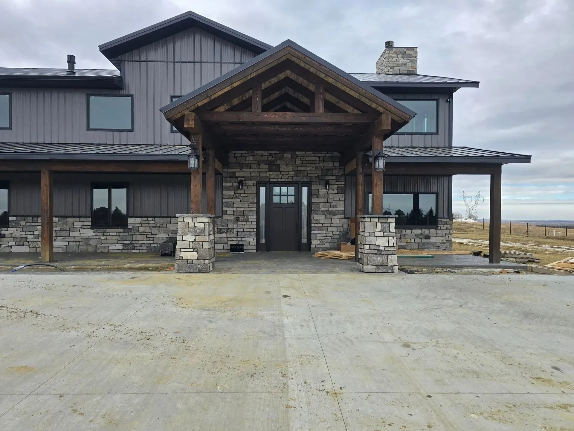 Two-story house with stone and wood accents. Porch over entrance, gray siding, concrete driveway, overcast sky.