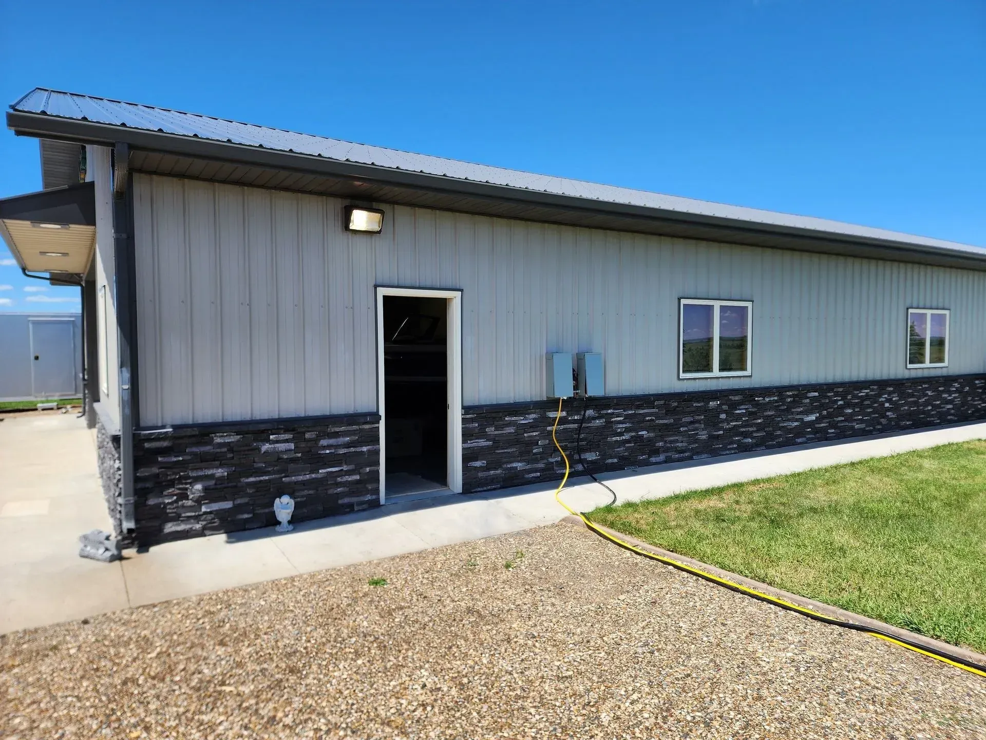 Exterior view of a gray building with a black stone facade, door open, windows, and a gravel driveway on a sunny day.