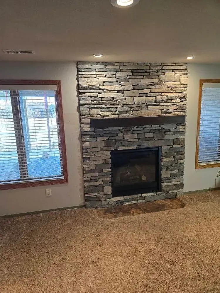 Fireplace with stacked stone facade and wooden mantel in a room with a carpeted floor and two windows.