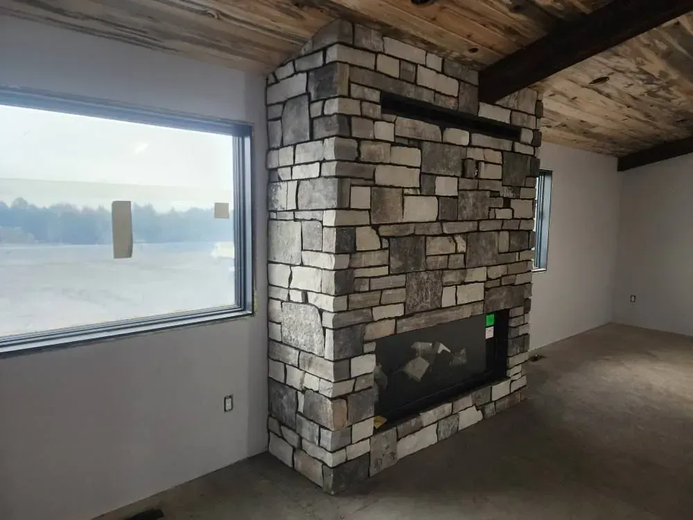 Stone fireplace with black inset in a room with a large window and wood ceiling.