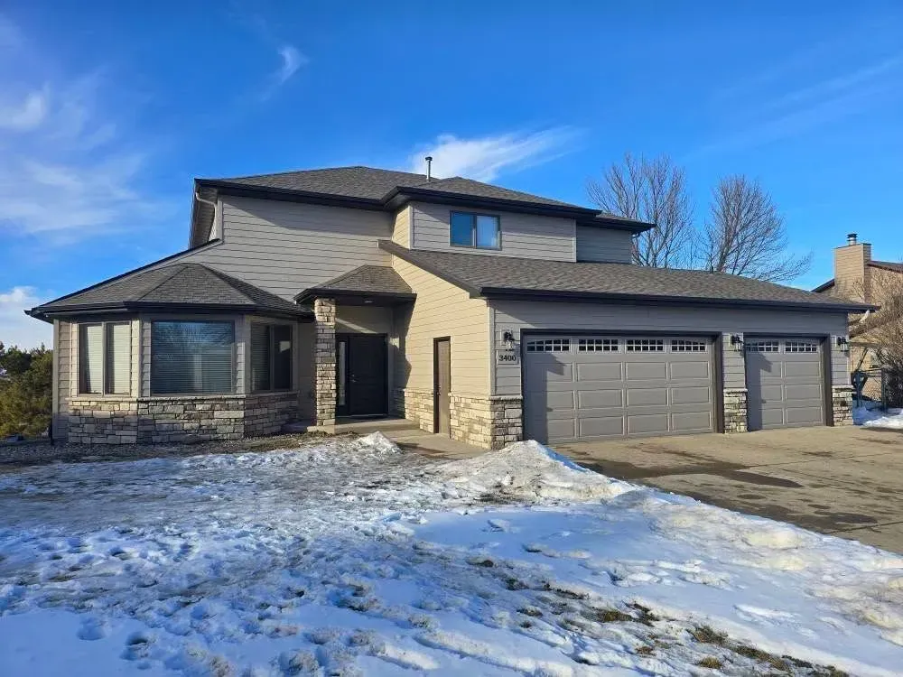 Two-story house with gray siding, stone accents, and three-car garage in a snowy setting.