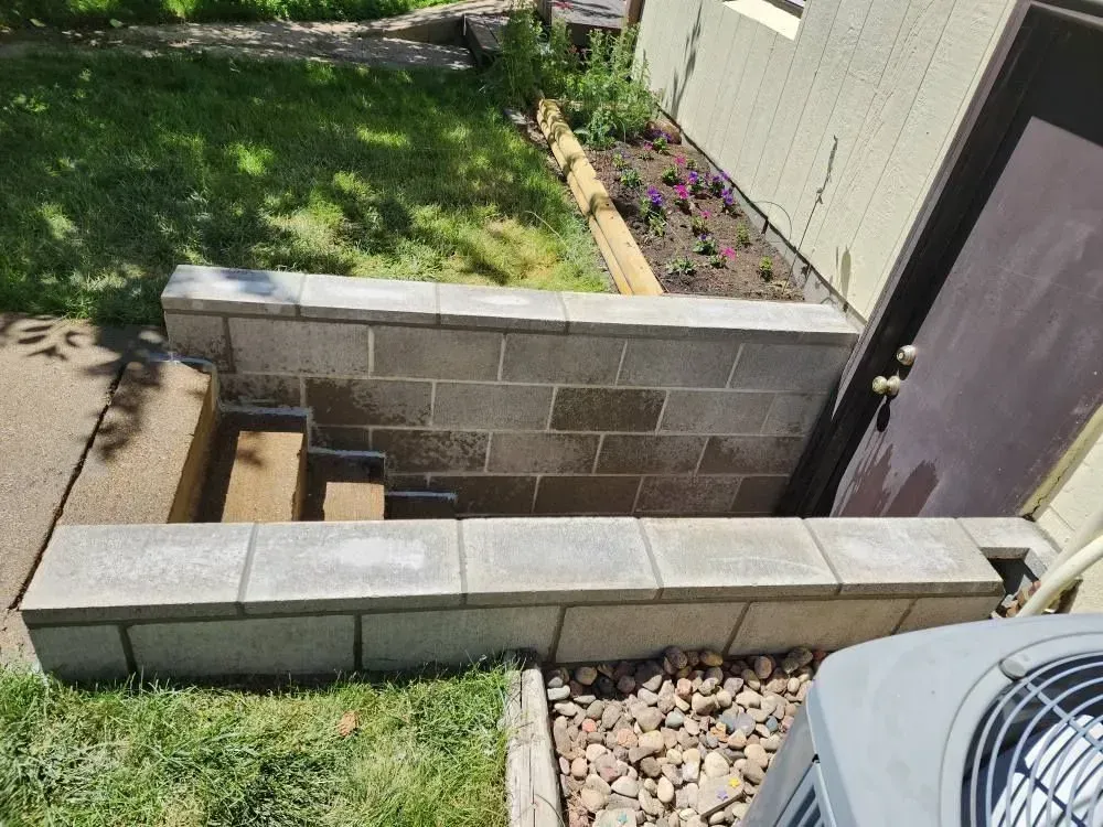 Concrete steps leading down into a basement entry. Gray block walls and trim. Small garden of flowers.