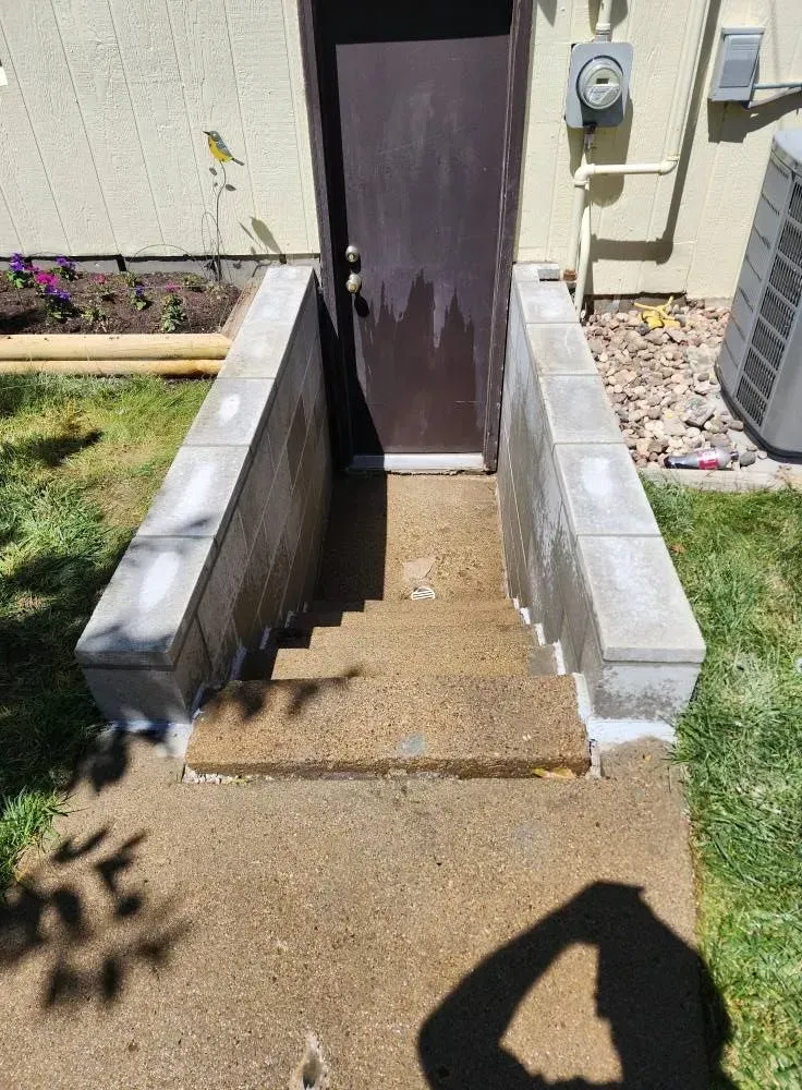 Concrete steps leading to a brown door with a gray cinder block retaining wall. Green grass and a flower bed are visible.