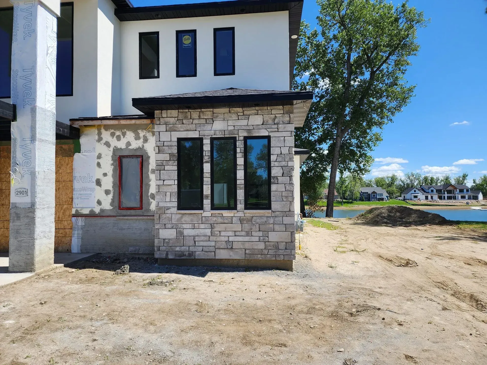 House under construction with stone facade and black framed windows, near a body of water.