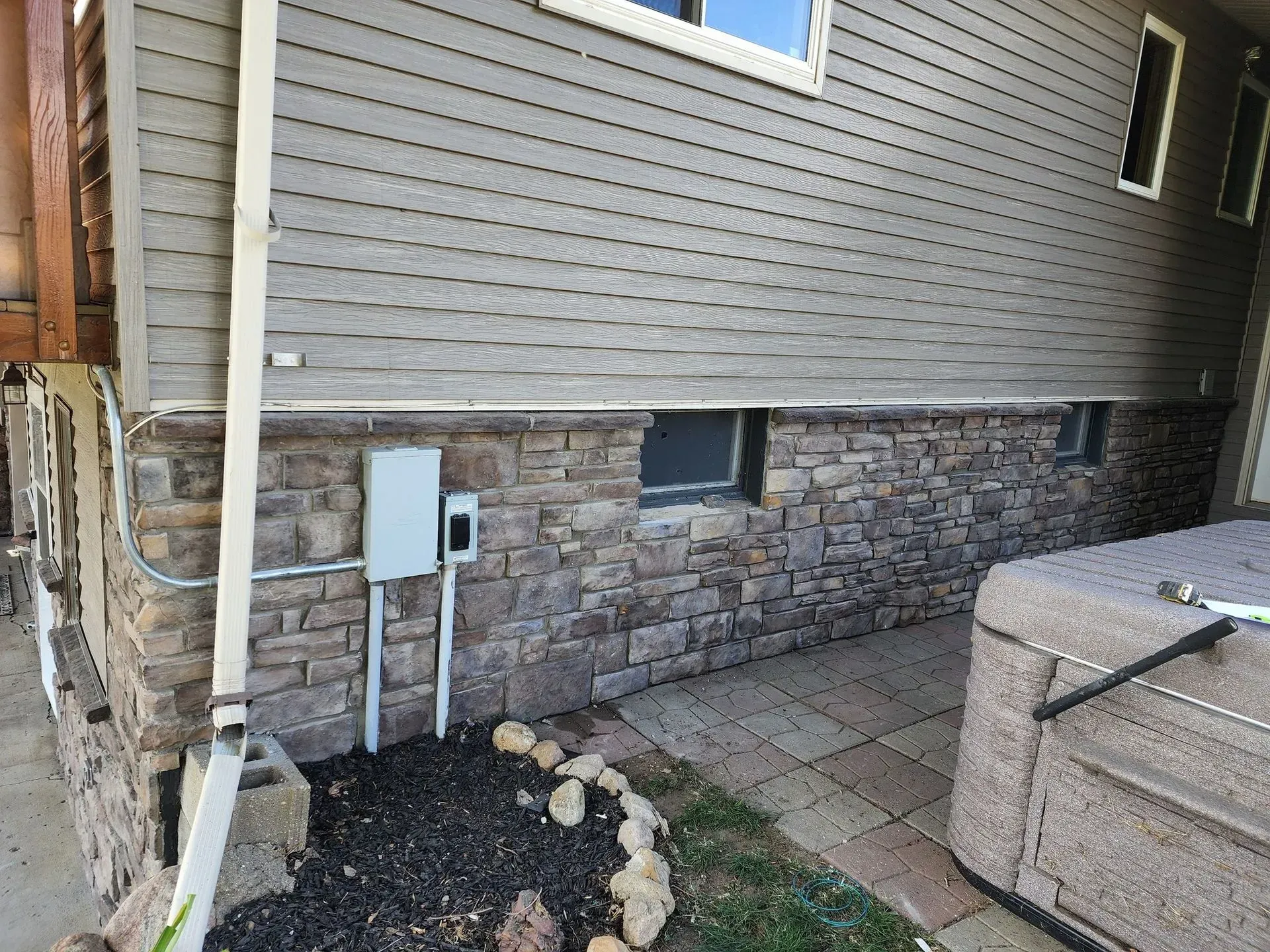 Exterior wall with stone base, gray siding, electrical box, small window, and hot tub.