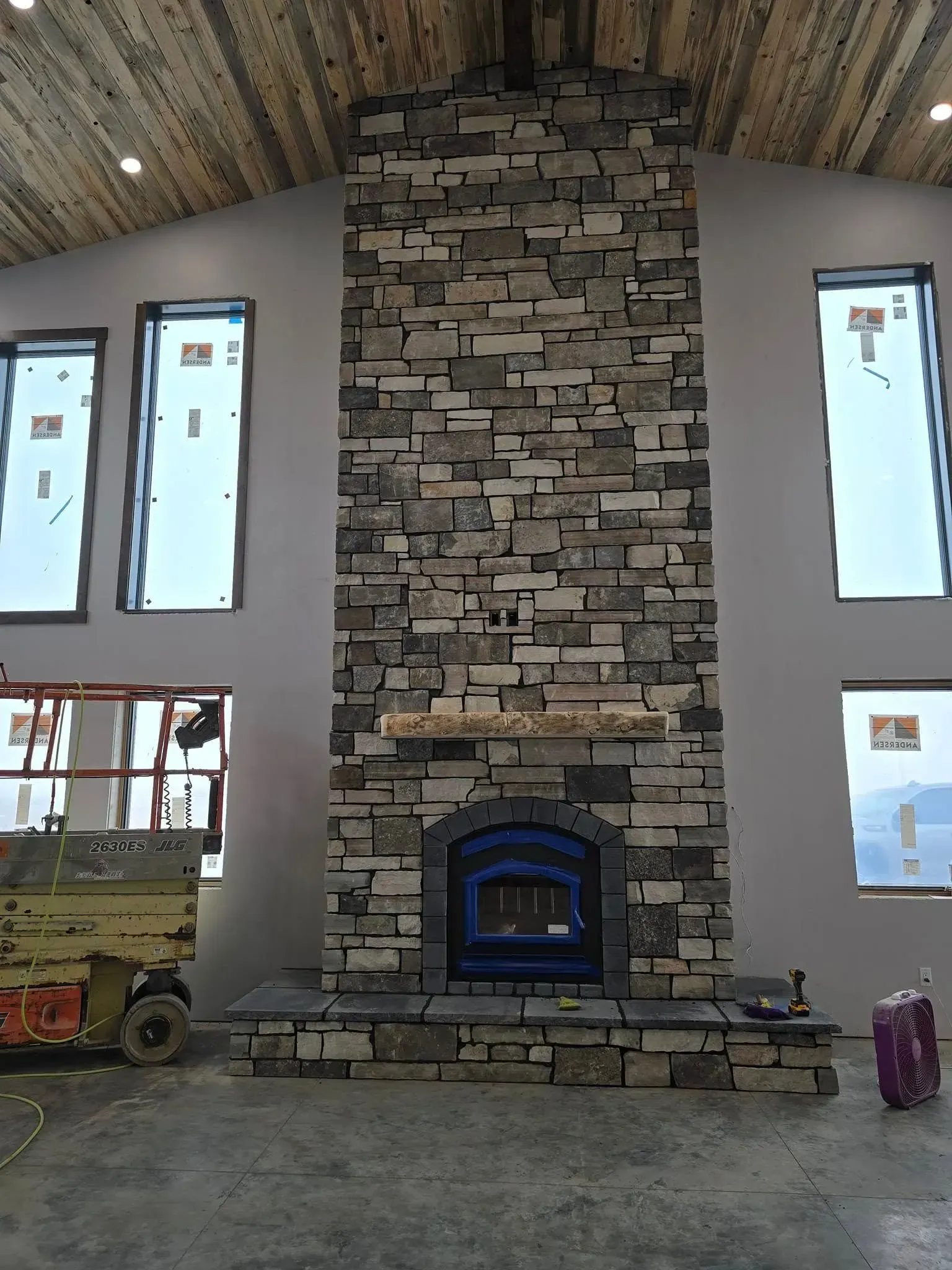 Stone fireplace in a room with large windows, unfinished construction.