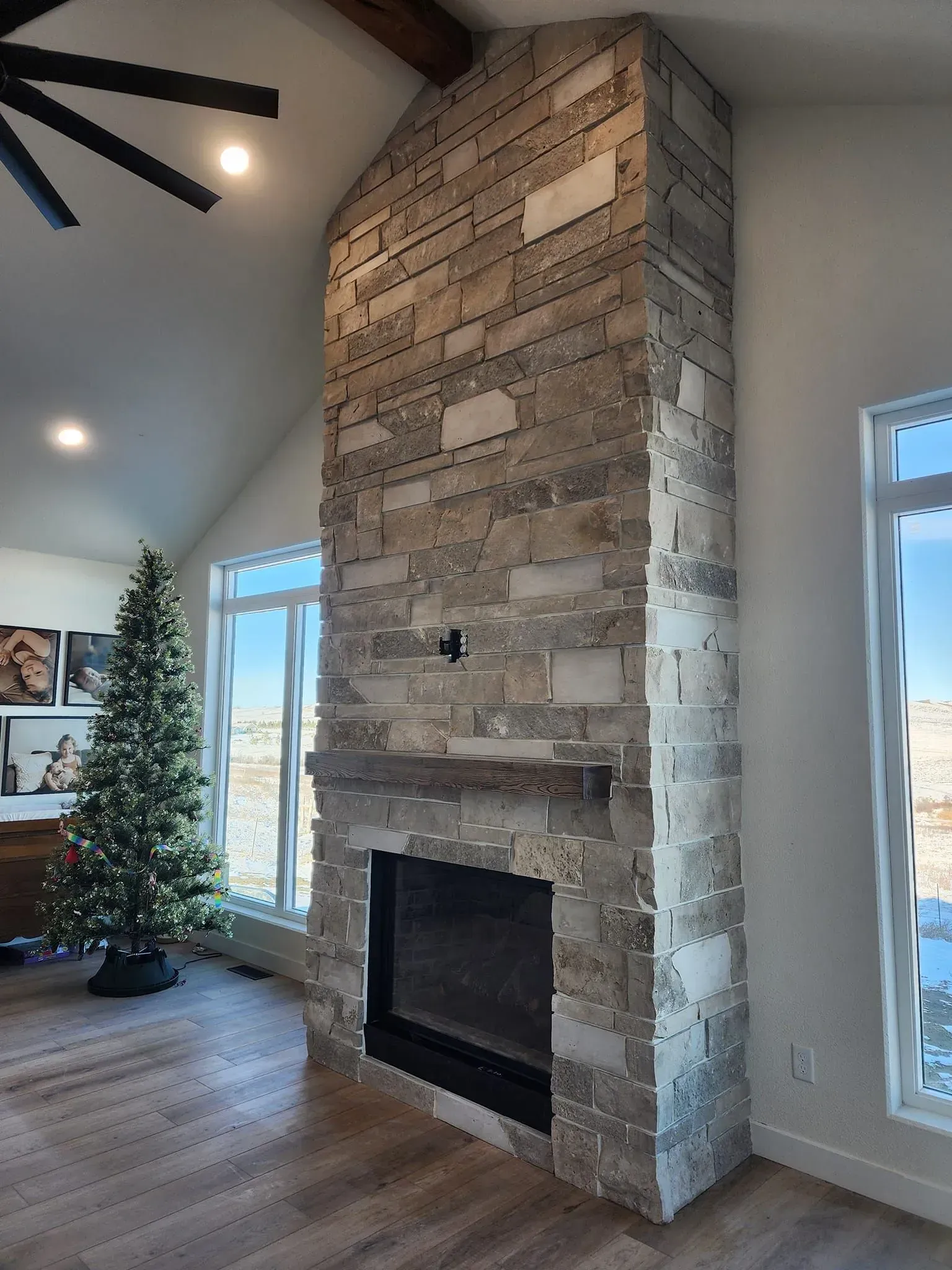 Stone fireplace with wooden mantle and high ceilings, next to large windows and a Christmas tree.