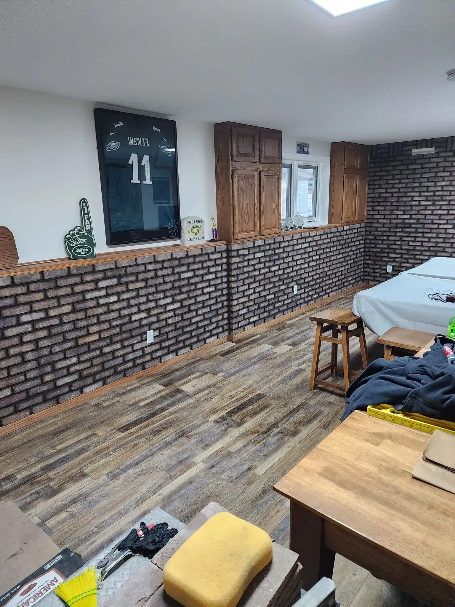 Basement room with faux brick wall, cabinets, wooden floor, jersey on wall, and small furniture.
