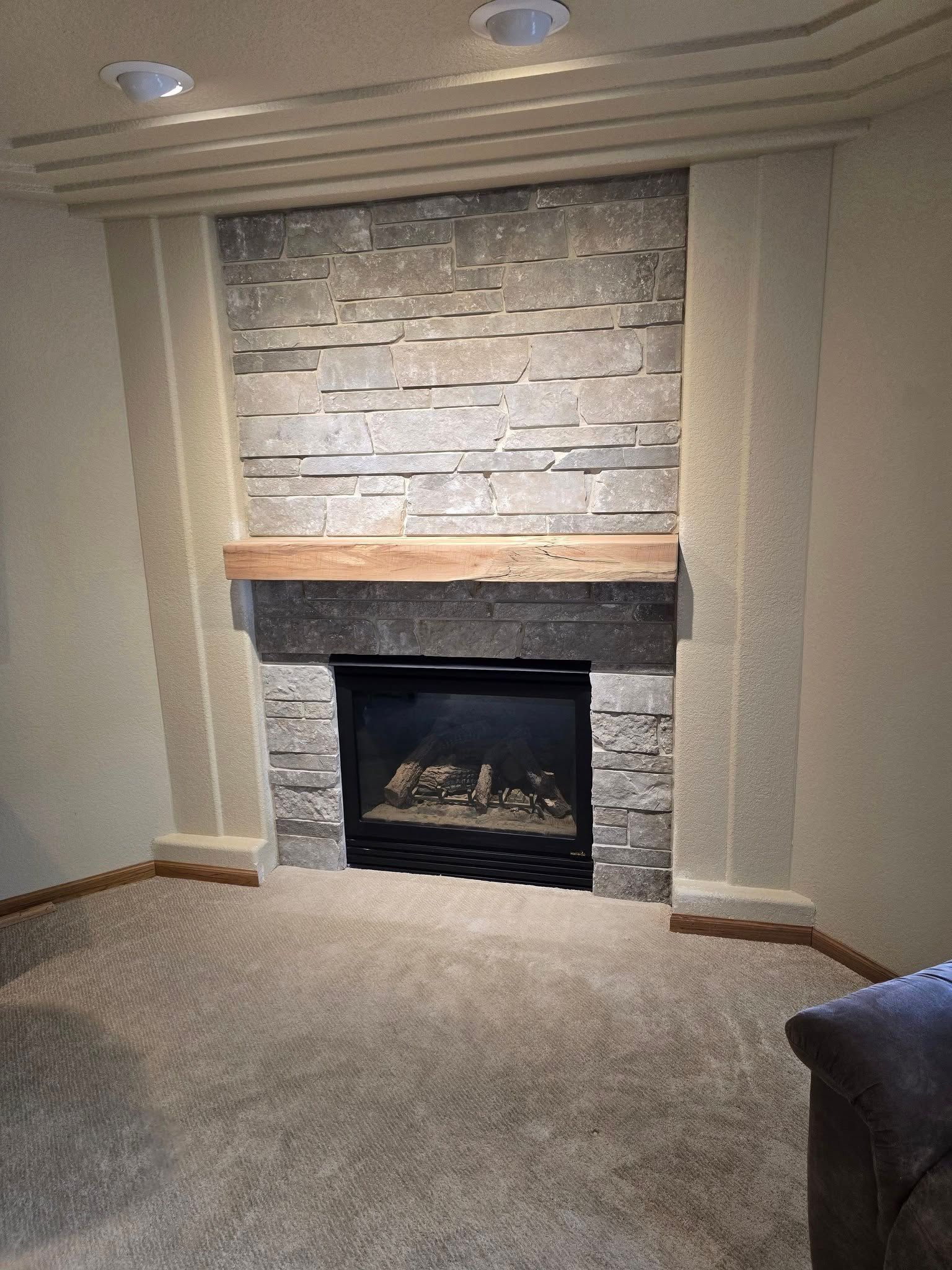 Fireplace with stone surround, wooden mantel, and built-in gas firebox, set in a room with tan walls and carpet.