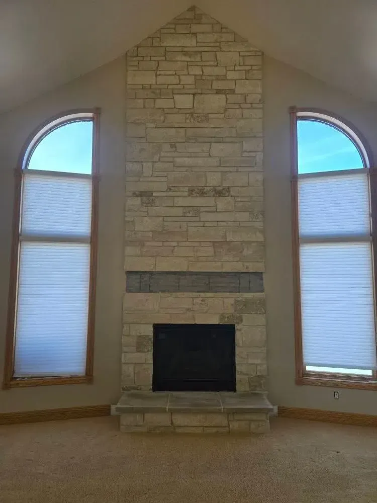 Fireplace with stone facade between arched windows, set in a light-colored room.