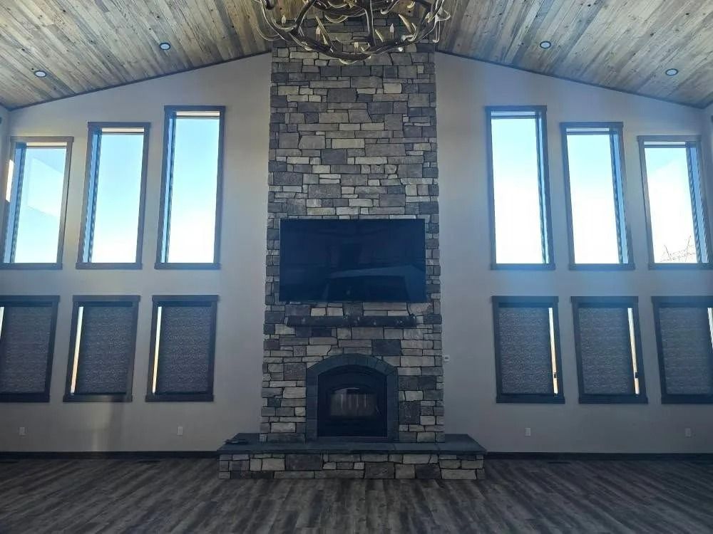 Room with stone fireplace, large windows, and a mounted TV. Wooden ceiling and floor.