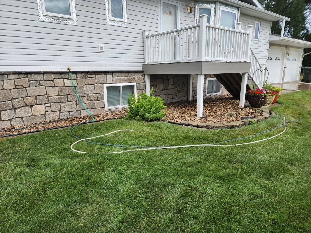 House exterior with a stone foundation, deck, and green lawn. A white hose lies on the grass.