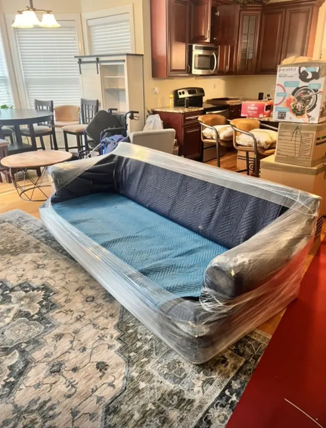 Wrapped blue sofa in a kitchen-living room, surrounded by furniture and boxes, suggesting a recent move.