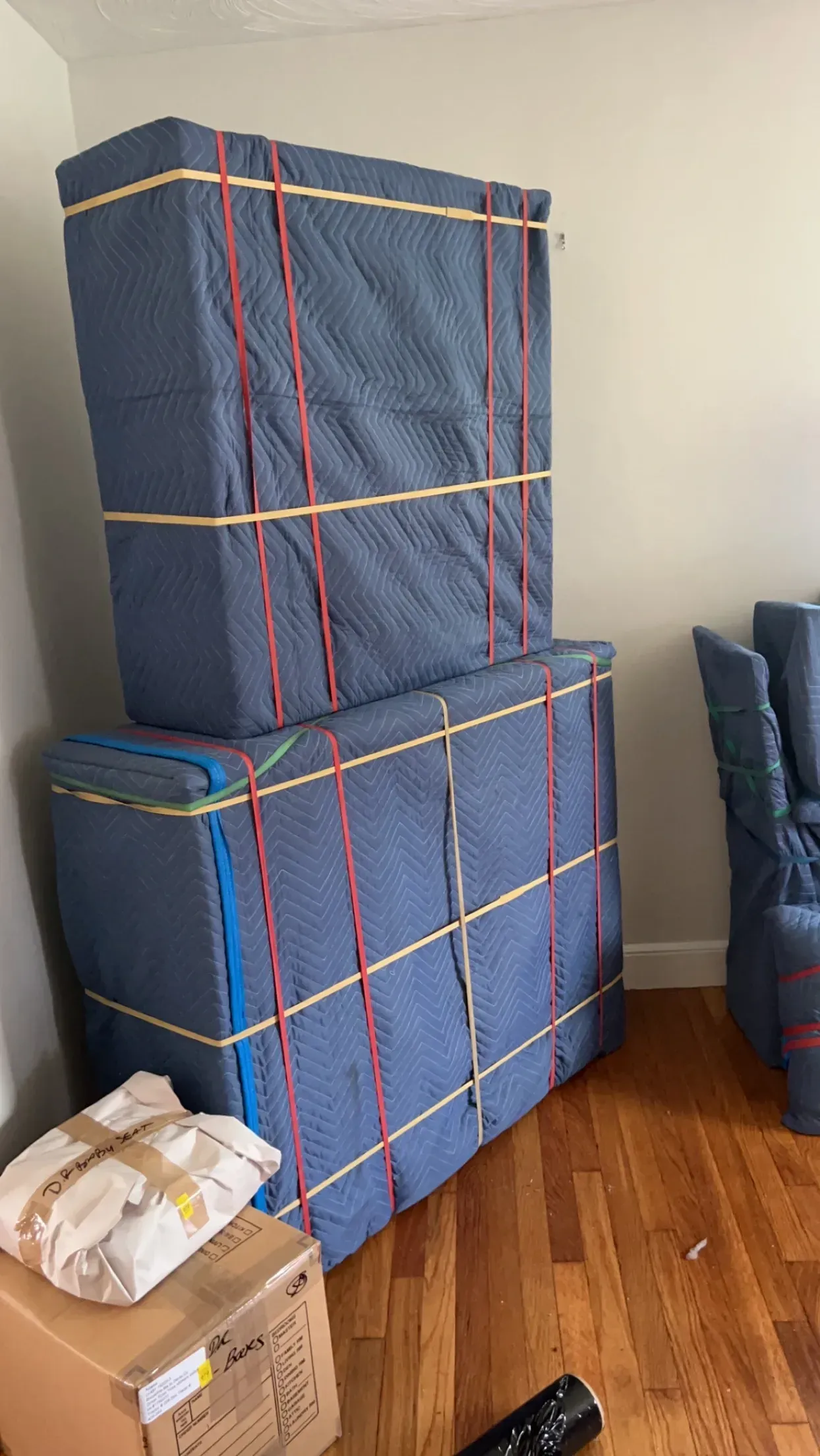 Stacked blue moving blankets tied with red straps in a room beside a cardboard box and chair