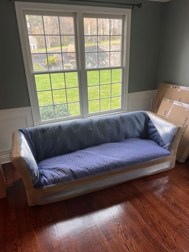 Blue-covered couch in a living room by a large window, with hardwood floors and cardboard boxes nearby