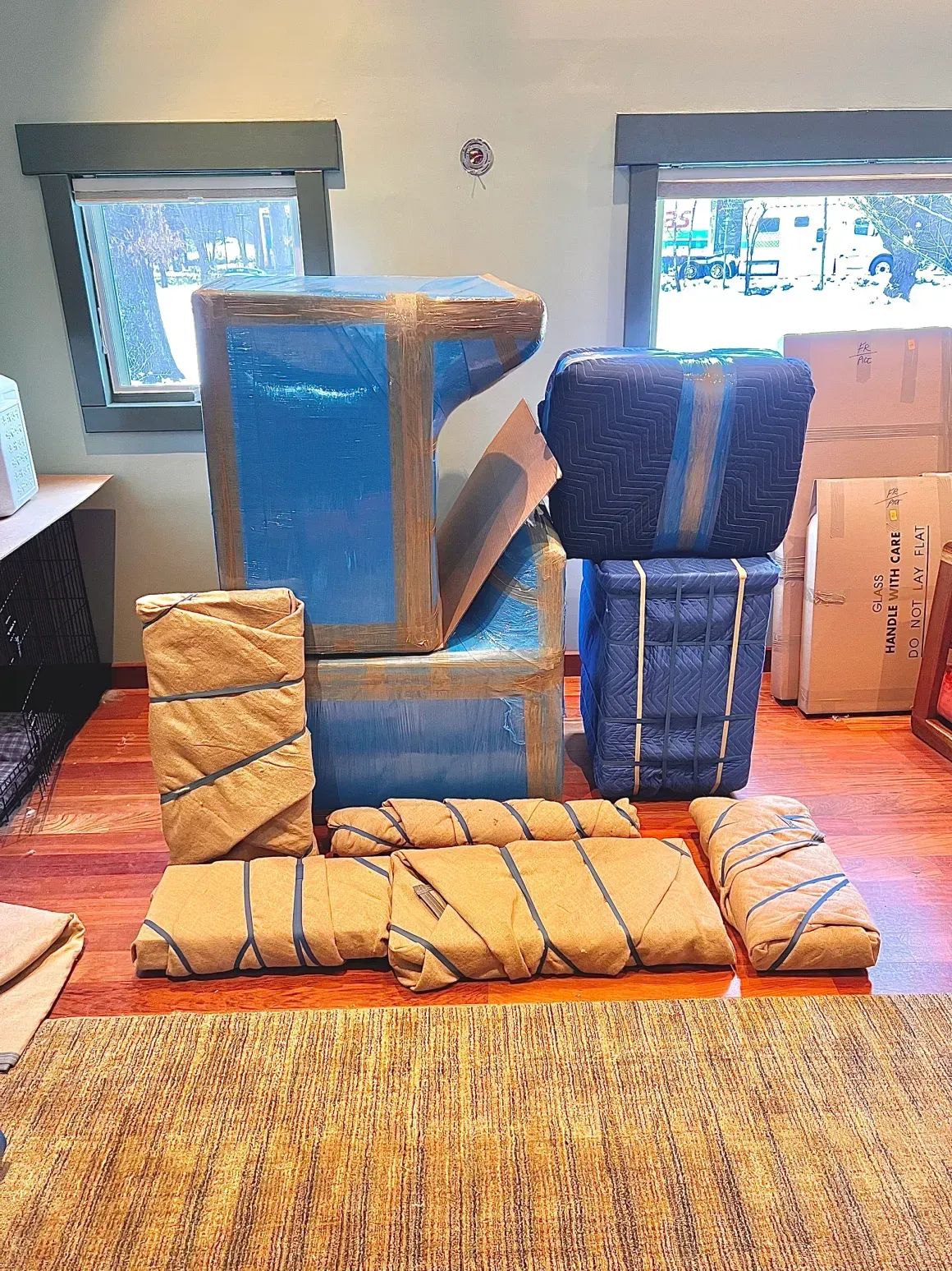 Packed furniture and boxes in a room with windows, wrapped for moving.