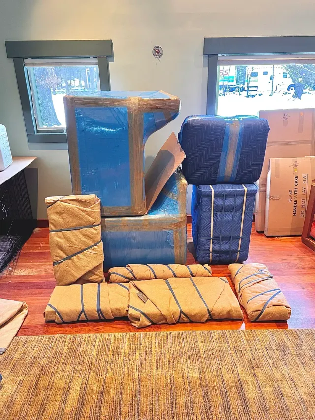 Stacked blue and brown packed furniture and boxes in a room with two windows and a rug on the floor