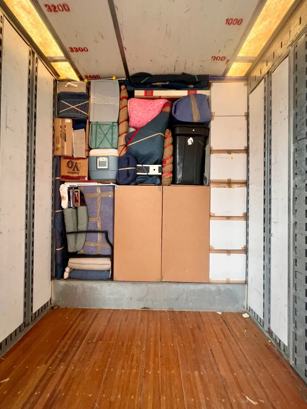 Inside a moving truck, stacked boxes, bags, and wrapped items fill the front wall behind an empty wooden floor.