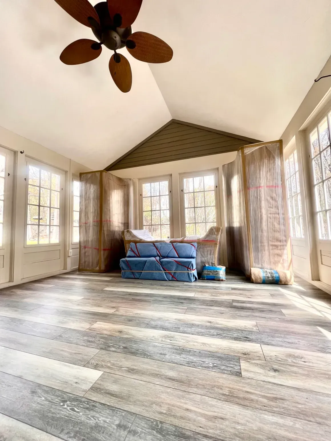 Empty sunlit room with large windows, wood floor, ceiling fan, and stacked blue cushions near the back wall