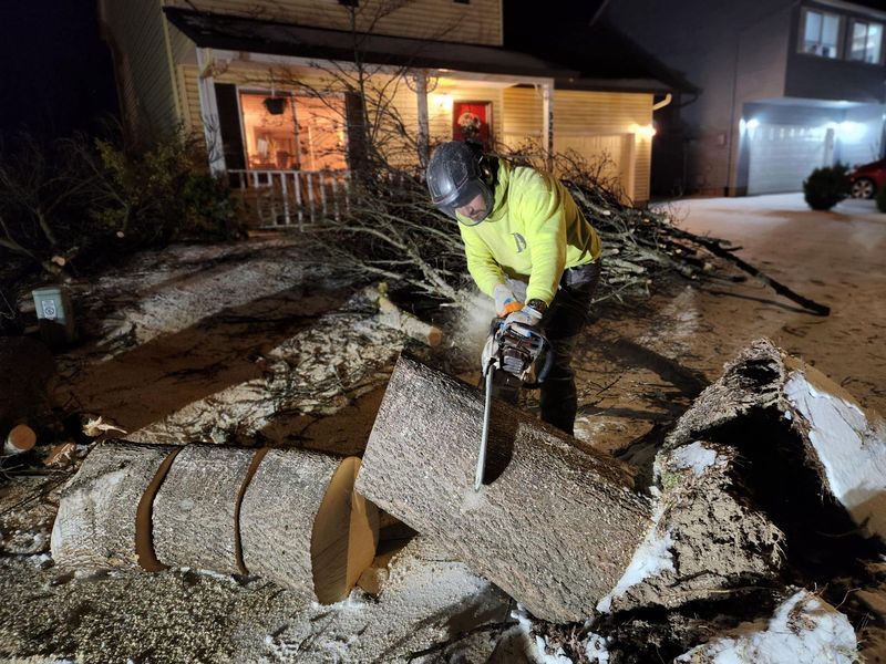 Person using chainsaw to cut a fallen tree in front of a house at night.
