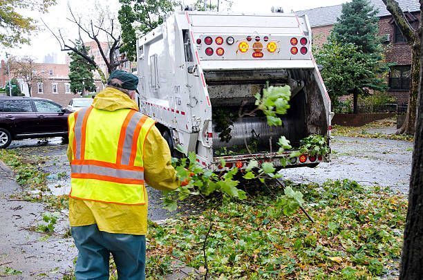 Garbage truck collecting green tree limbs, worker in yellow rain gear on street.