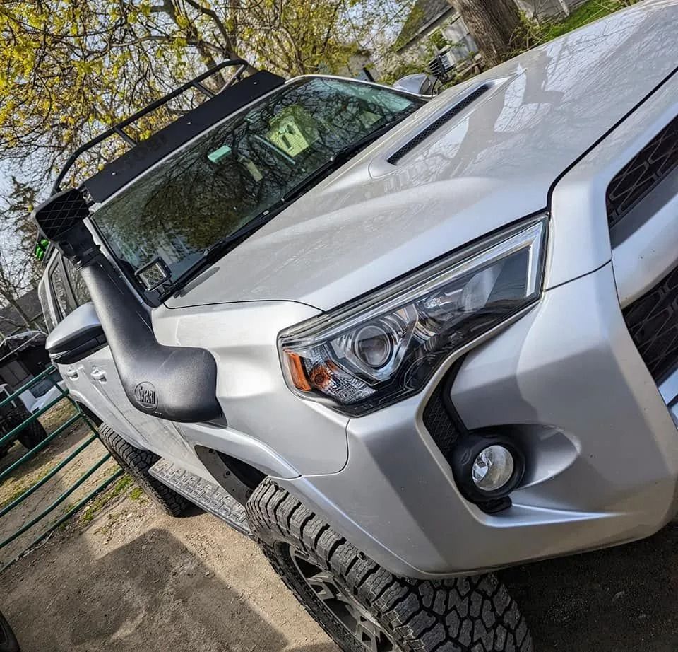 Silver off-road SUV parked at an angle on a dirt driveway, front end visible and roof rack attached