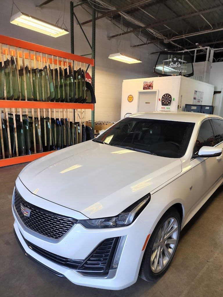 A white cadillac is parked in a garage next to a rack of windshields.