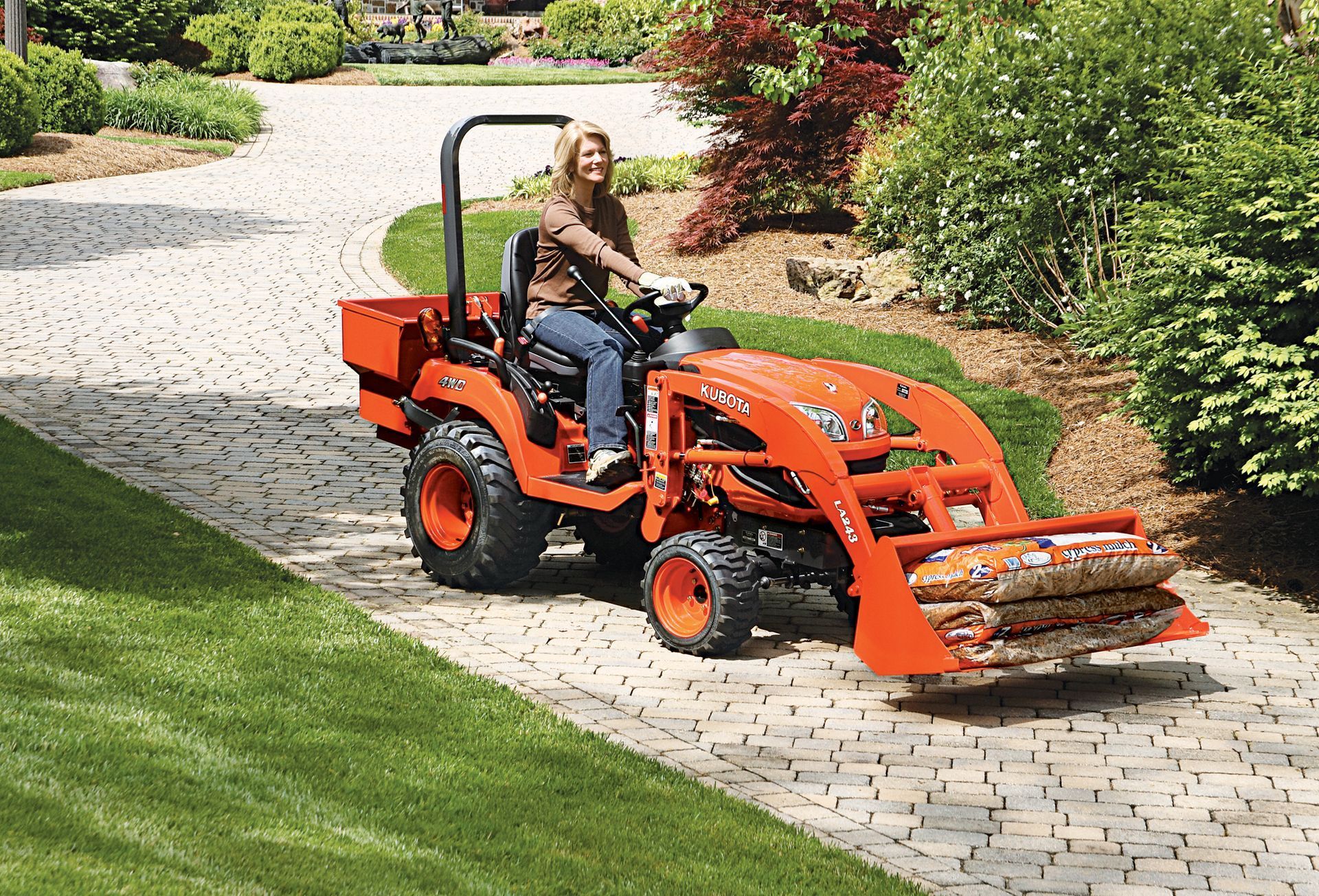 Woman operating an orange tractor on a paved driveway, carrying bags of material in the front bucket.