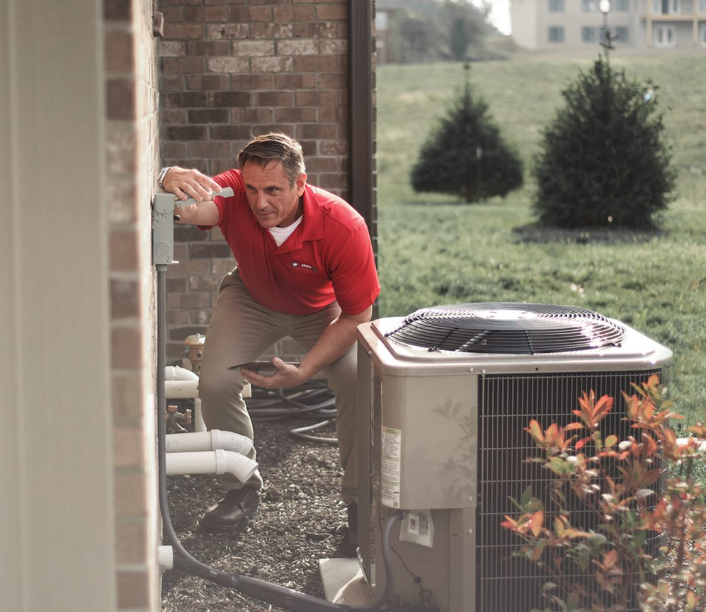 HVAC technician inspecting an air conditioning unit outdoors, holding tablet, near brick wall, in daylight.