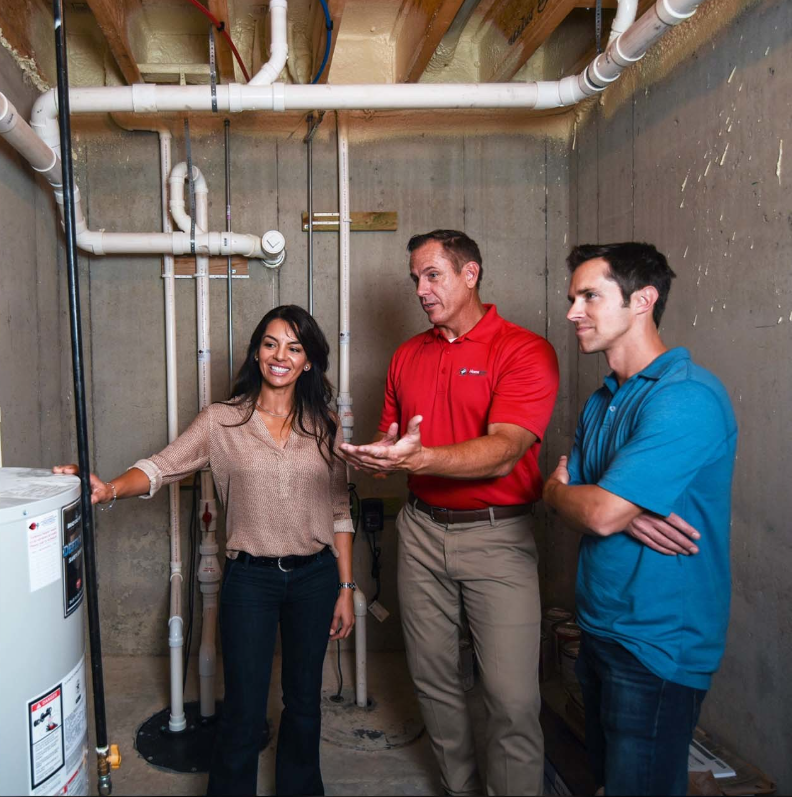Three people in a basement inspecting a water heater; the man in red gestures.
