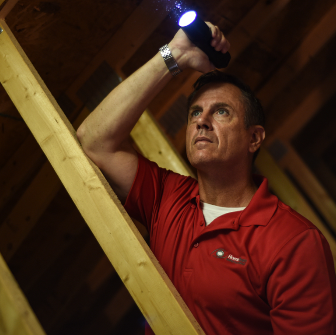 Man in red shirt in attic, holding flashlight, inspecting overhead.