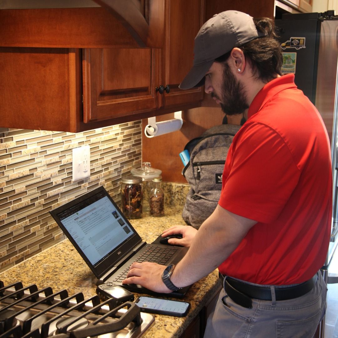 Man in red shirt and gray hat working on a laptop at a kitchen counter.