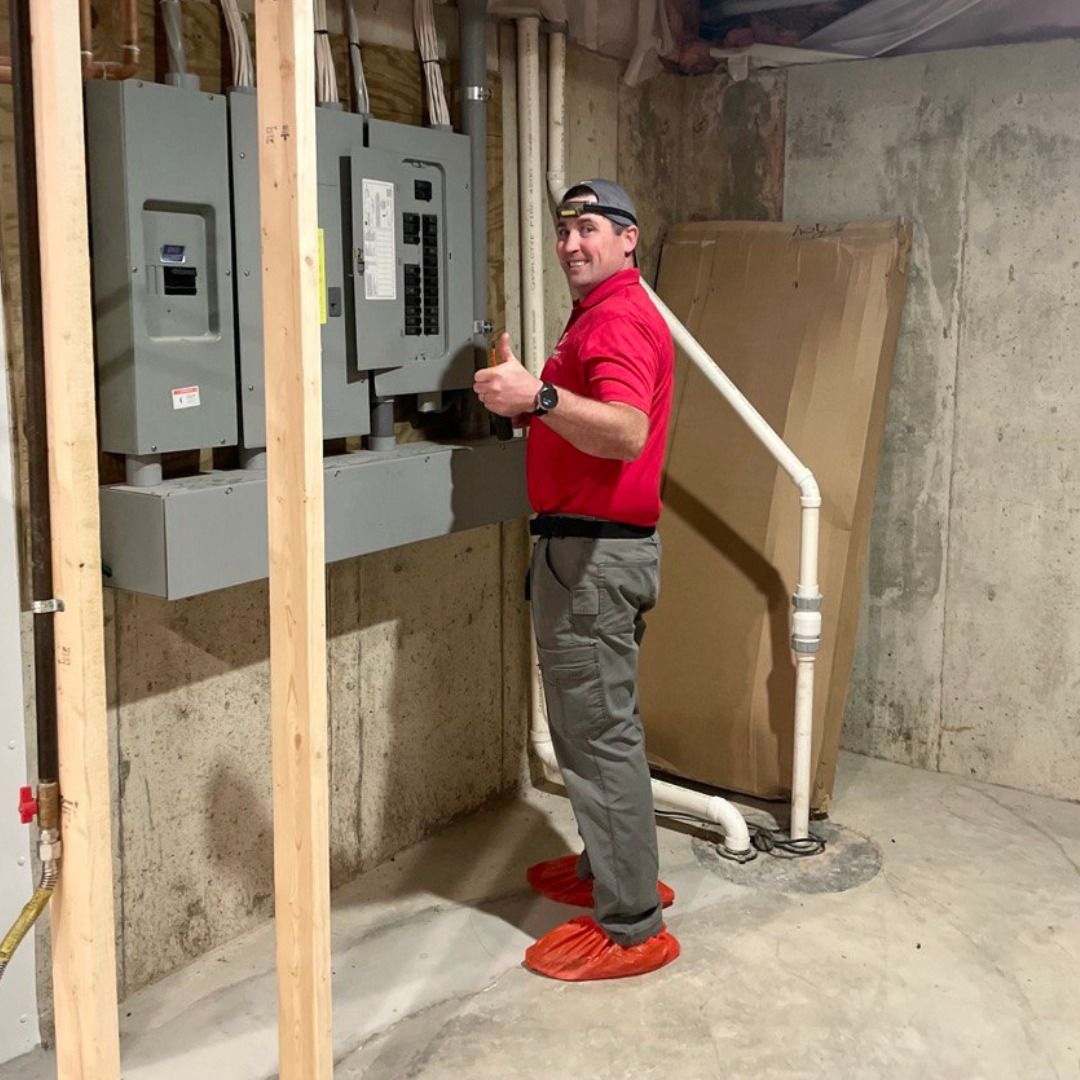 Man in red shirt and gray pants gives thumbs up in a basement with electrical panels. He is wearing orange shoe covers.