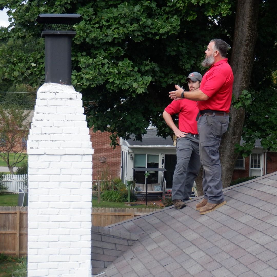 Two men on a roof inspecting a chimney, both wearing red shirts, next to a painted brick chimney.