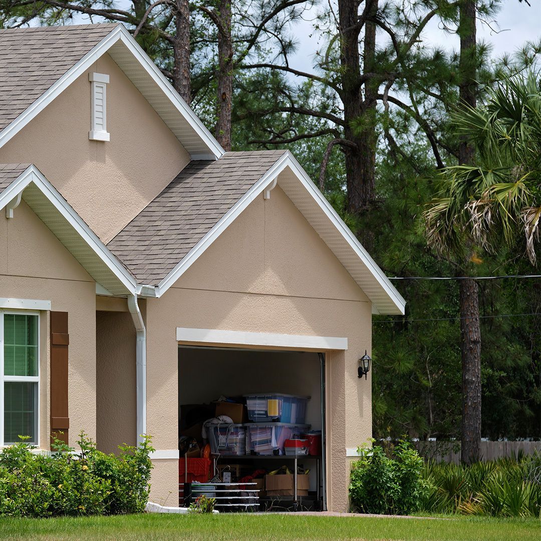 Tan house with open garage filled with boxes, with green grass and trees in the background.
