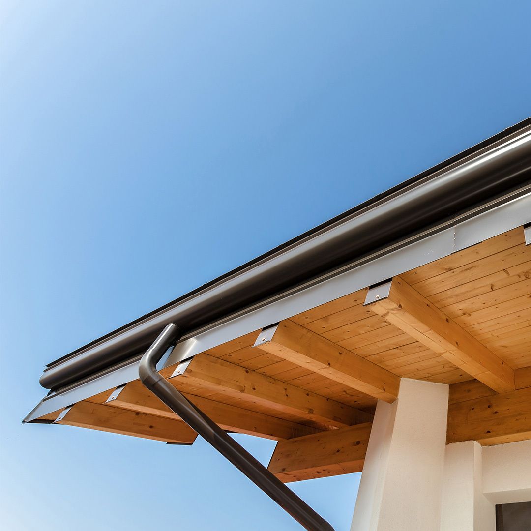 Brown gutters and downspout on wooden eaves against a bright blue sky.