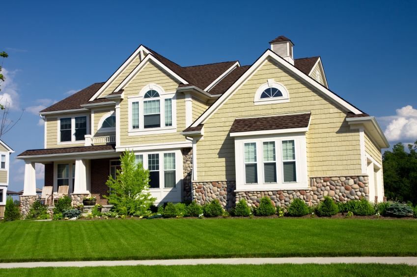 Two-story house with tan siding, stone accents, and a well-manicured lawn under a blue sky.