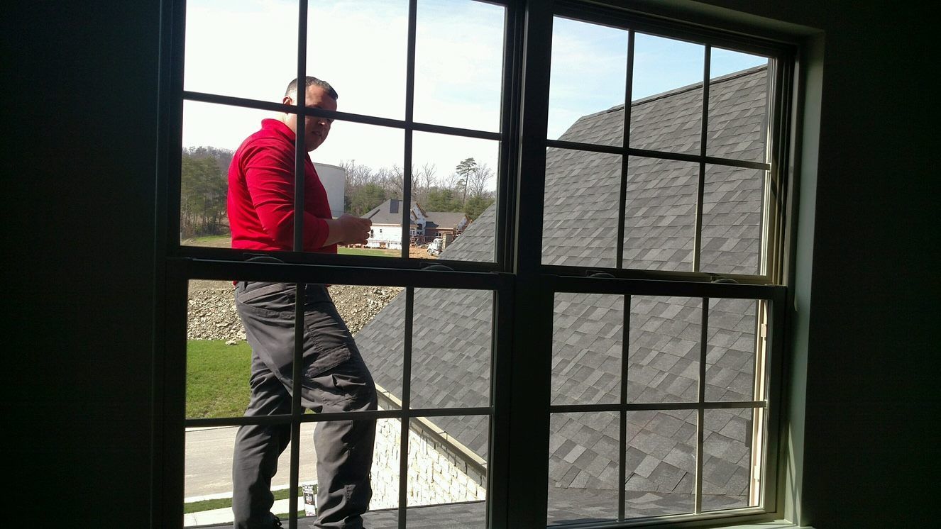 Man in red shirt cleaning window from outside, against roof and sky.